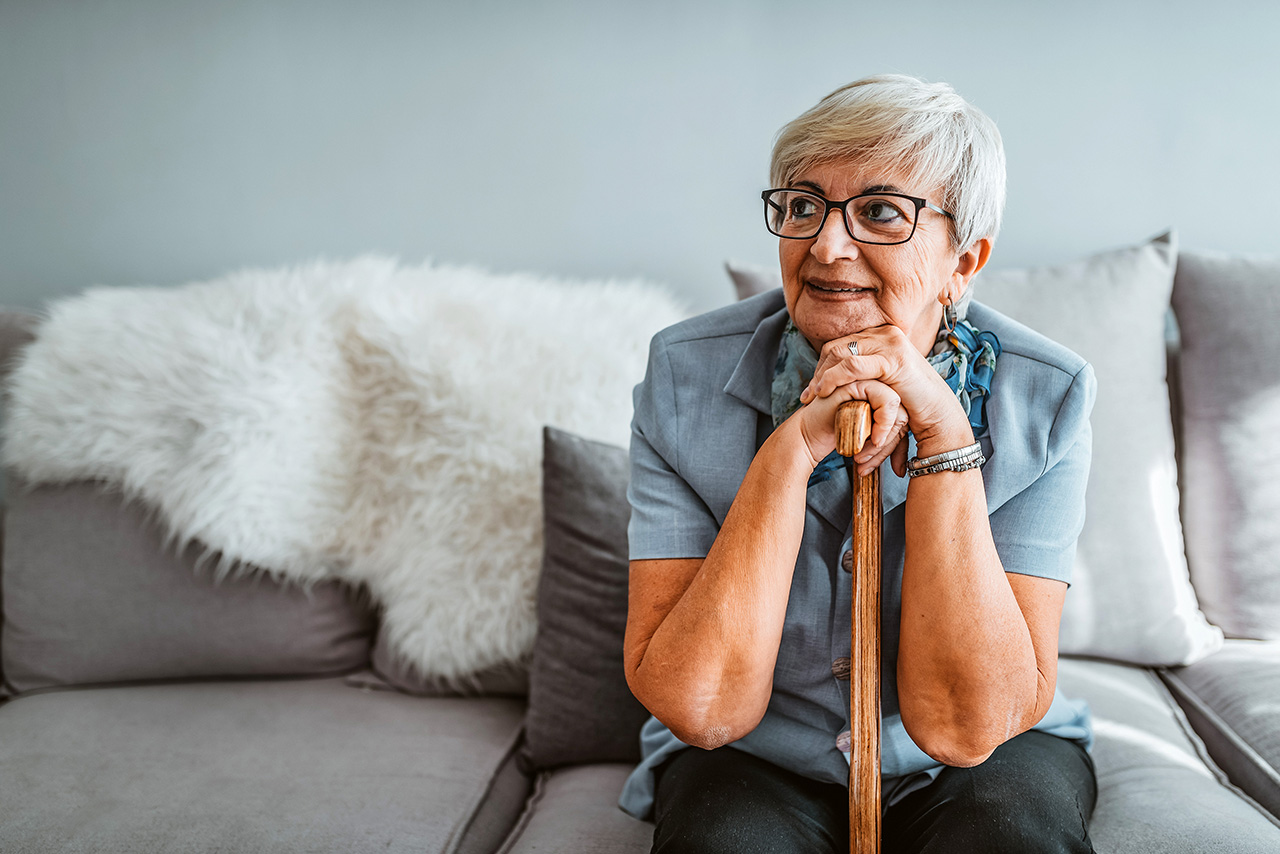 Ältere Frau mit kurzen grauen Haaren, die zu Hause auf dem Sofa sitzt, gestützt auf einem Gehstock.