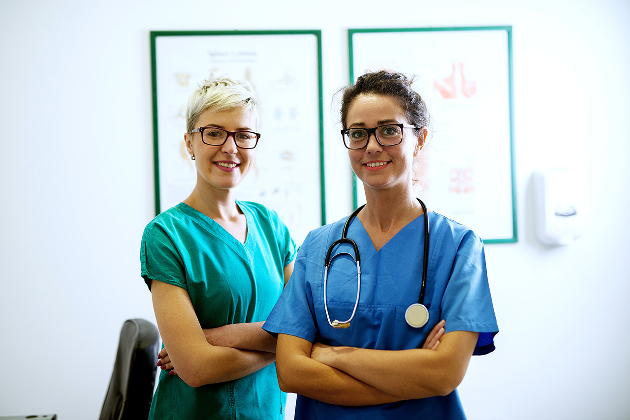 Porträt von zwei Krankenschwestern mit Brille, die mit verschränkten Armen im Büro stehen und in die Kamera lächeln.
